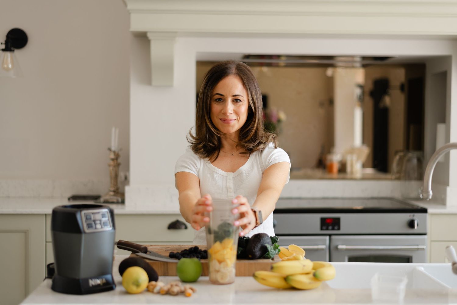 Someone stands in a bright white kitchen preparing food with fresh ingredients and a blender on the counter.