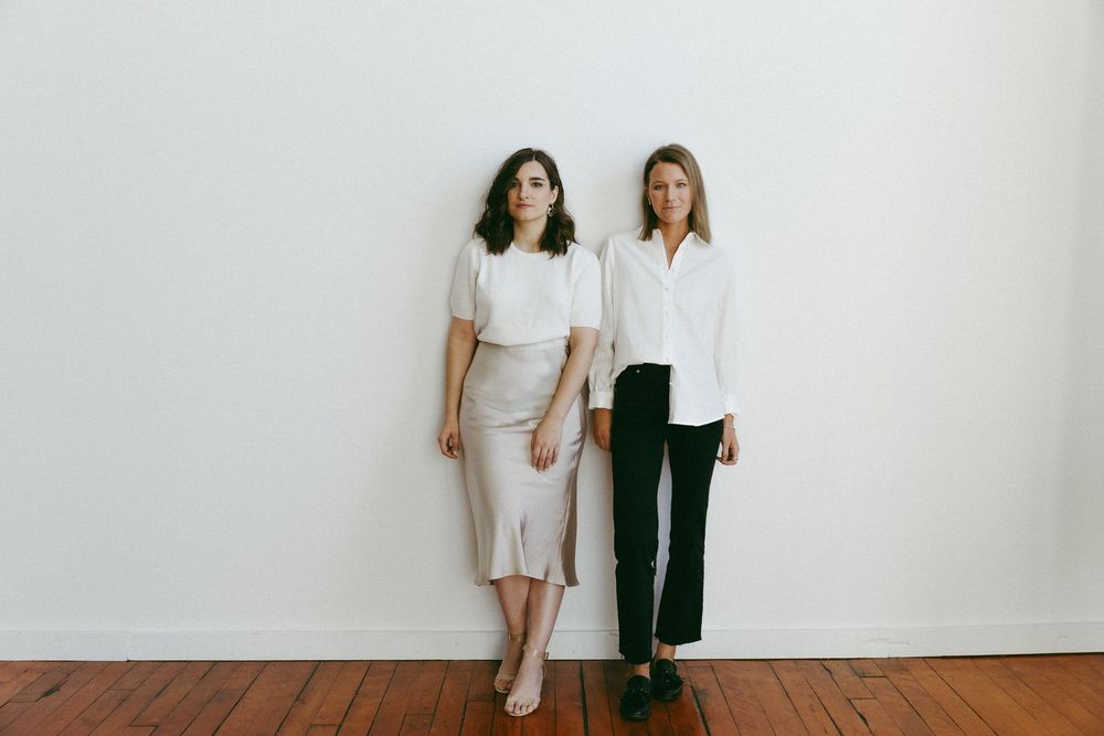 Two people stand together near a white wall showcasing minimal monochromatic outfits and relaxed styling.