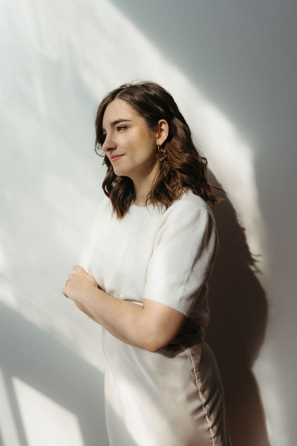 A woman in a white t-shirt poses against a wall with dramatic shadows casting geometric patterns.