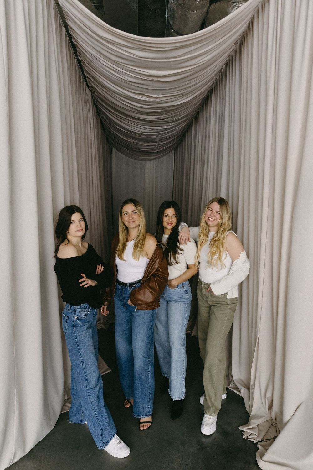 Group of people wearing jeans standing against white draped curtains in artistic photo composition.