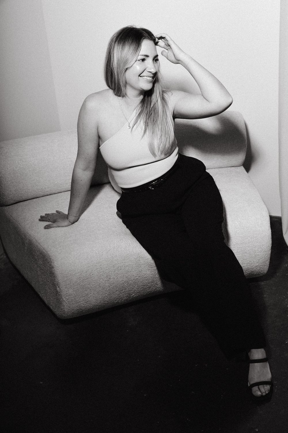 Black and white portrait of a smiling person in a crop top and dark pants lounging on a light colored seat.