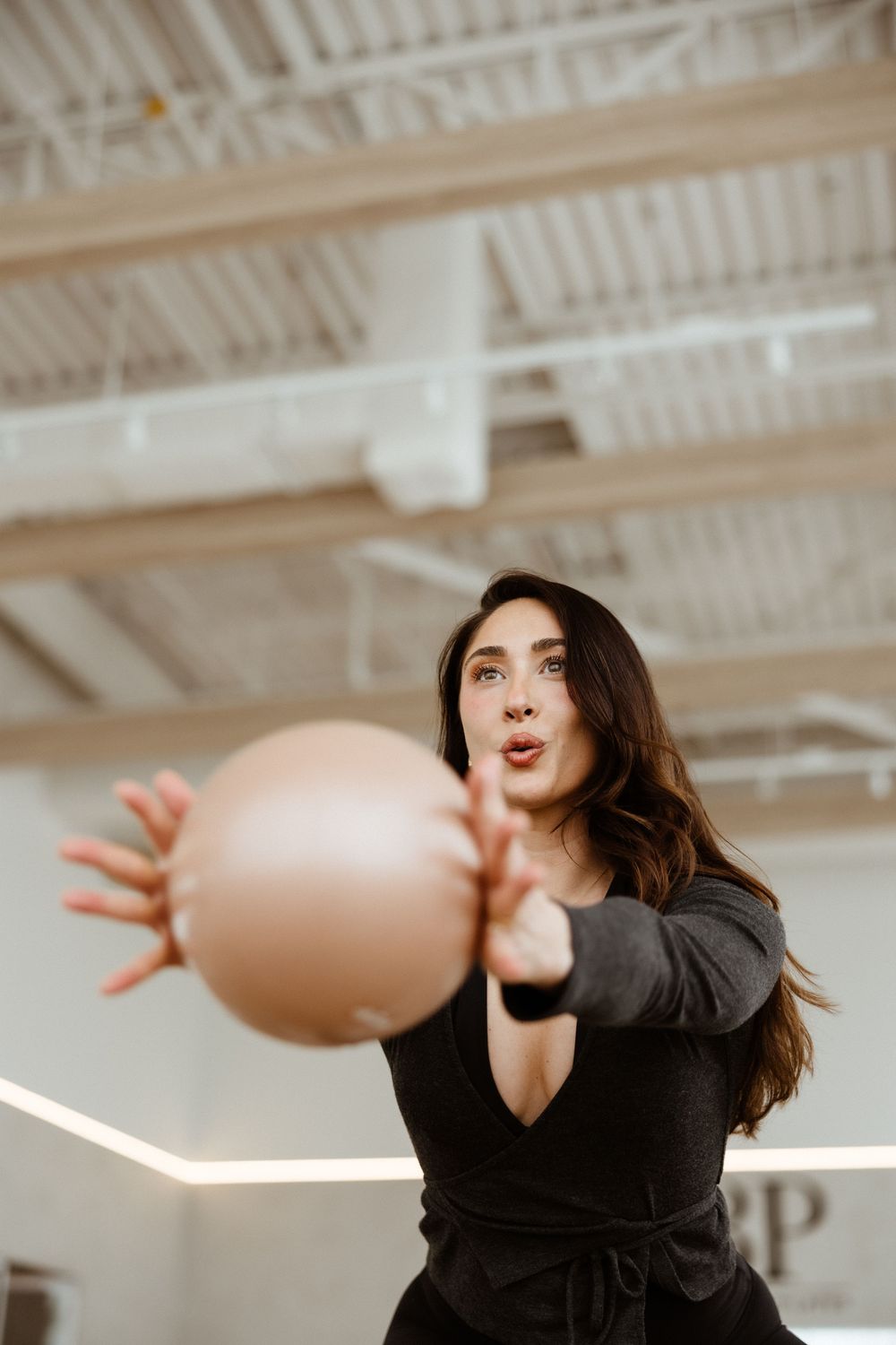 Someone in a black dress serves a volleyball at an indoor gym with white industrial ceiling.