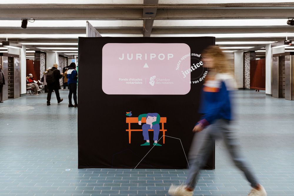 A blurred pedestrian walks past a black wall with a pink sign in an underground subway station corridor.