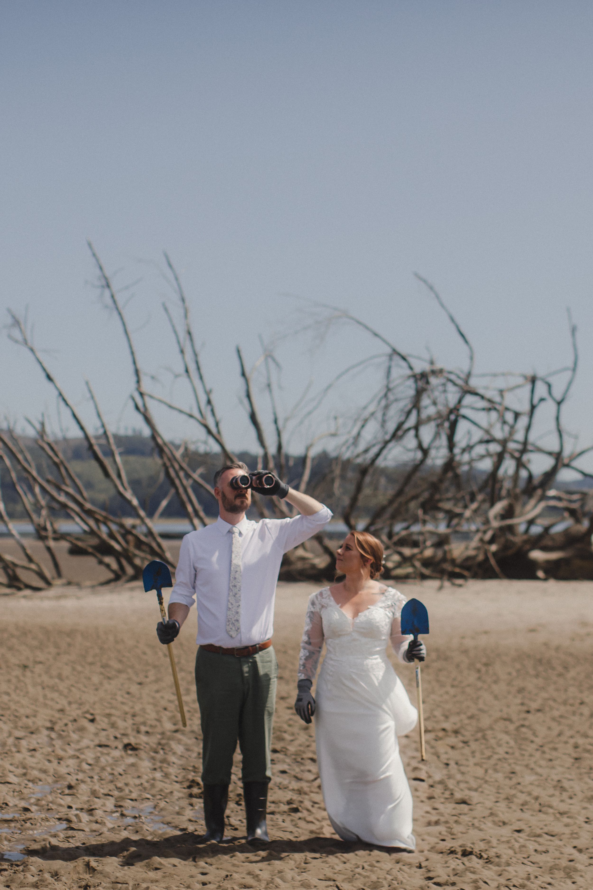 Wedding couple poses playfully on driftwood-covered beach under cloudy skies.