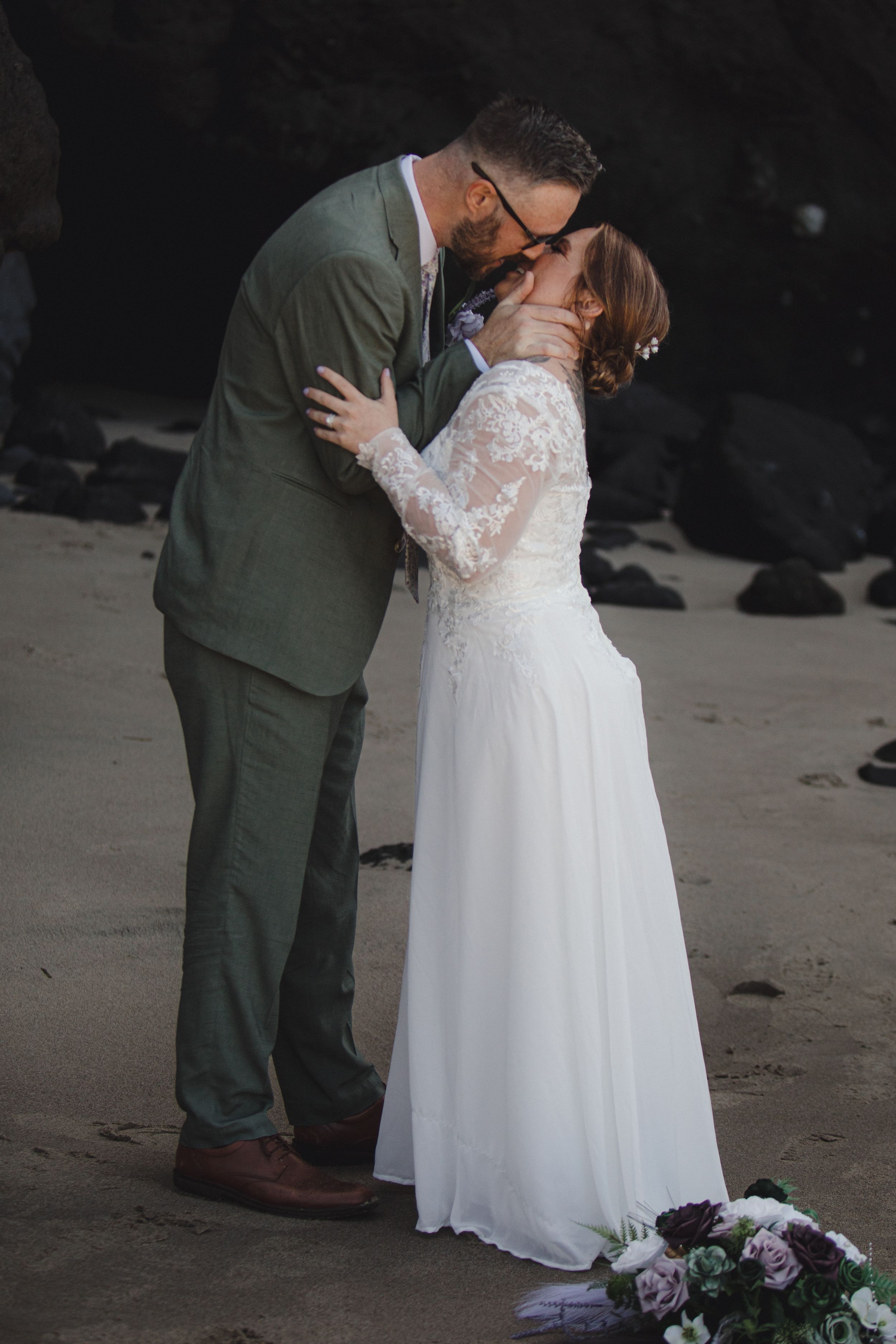 Tender kiss between newlyweds on a moody beach with florals at their feet.