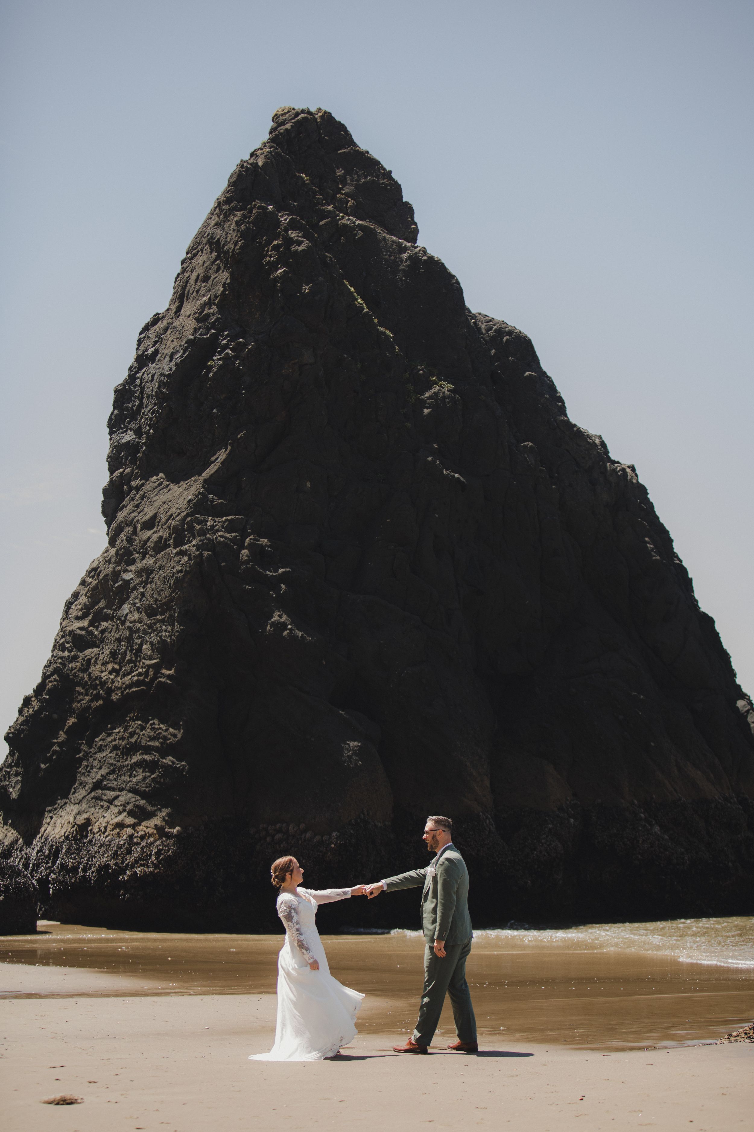 A couple in wedding attire share a romantic moment on a beach with a dramatic rock formation in the background.
