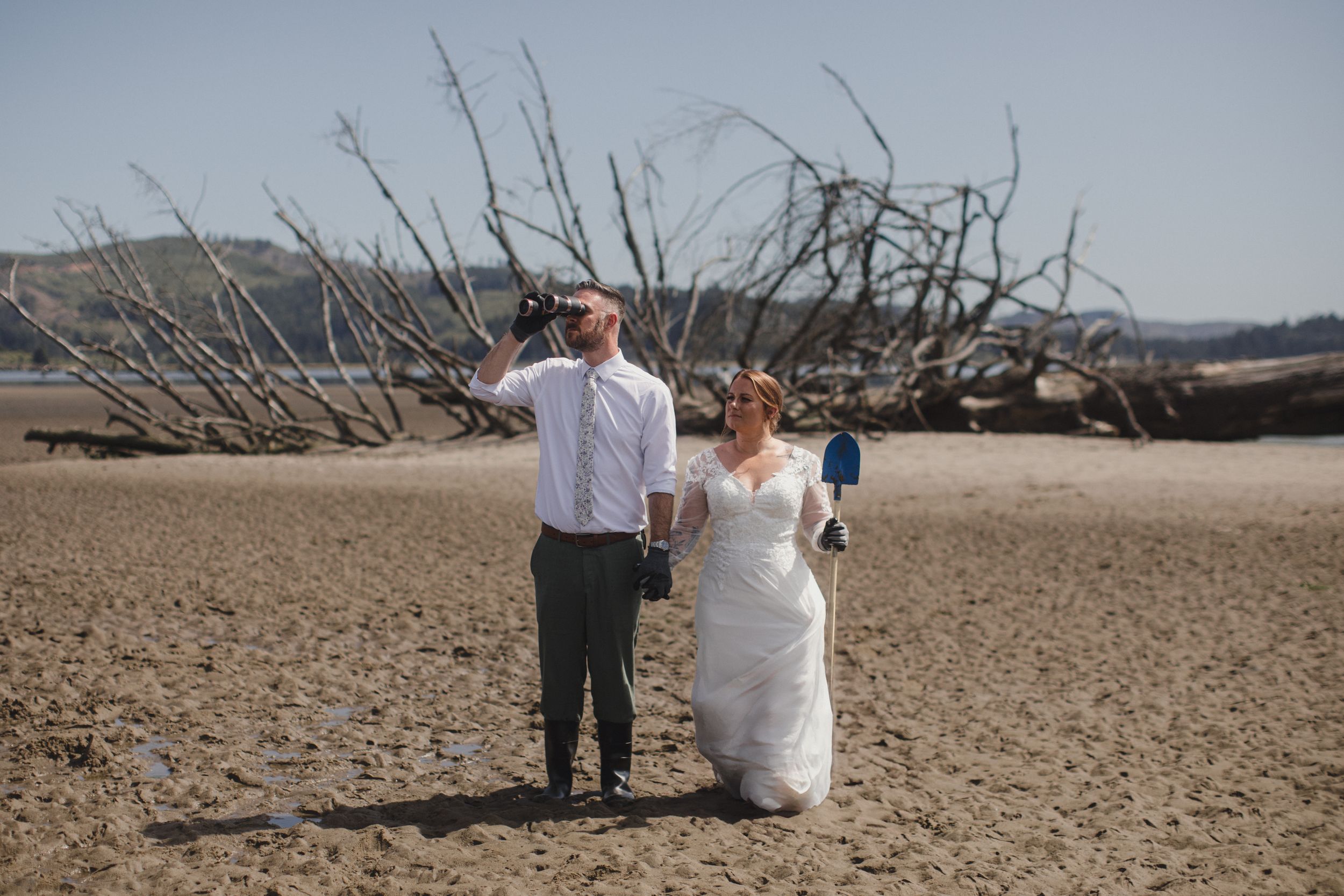 A couple poses in wedding attire on a beach with driftwood scattered in the background.