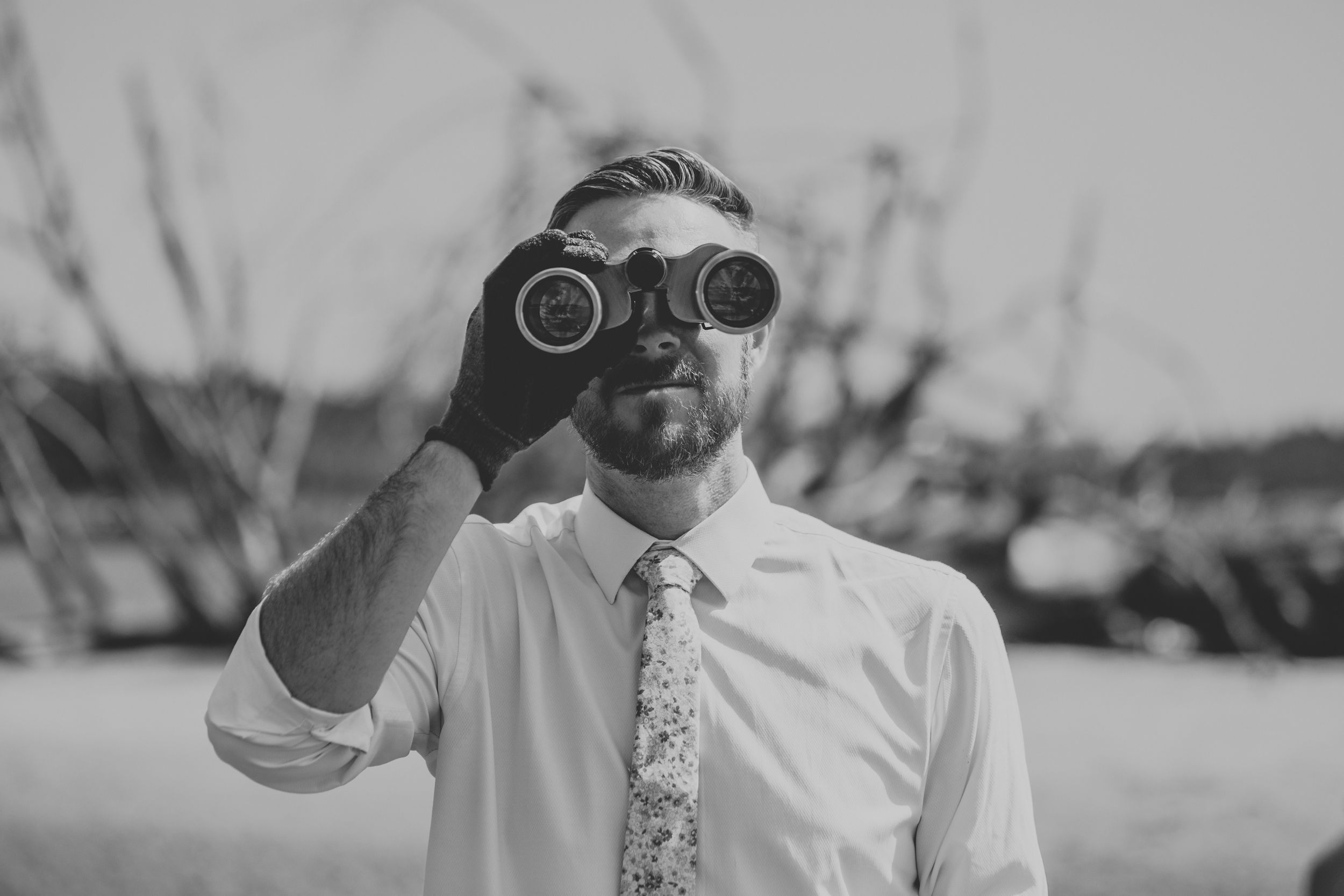 Someone wearing a white dress shirt and floral tie looks through binoculars in black and white.