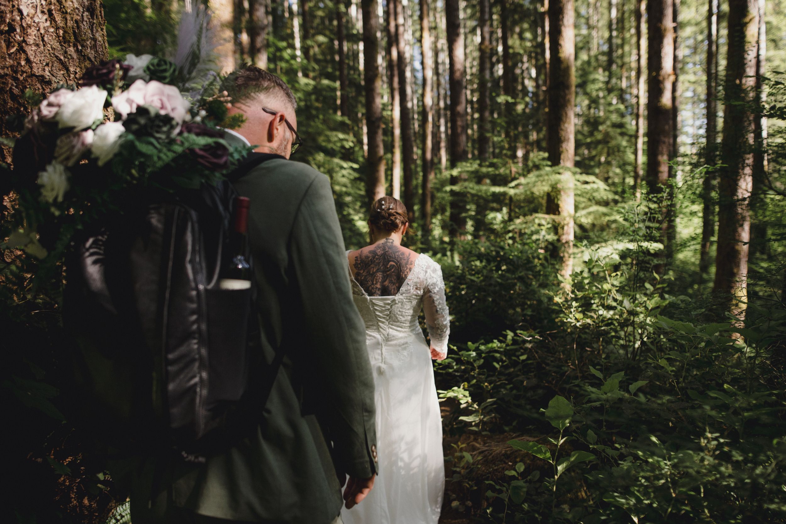 Wedding couple walks through tall forest with white floral bouquet.