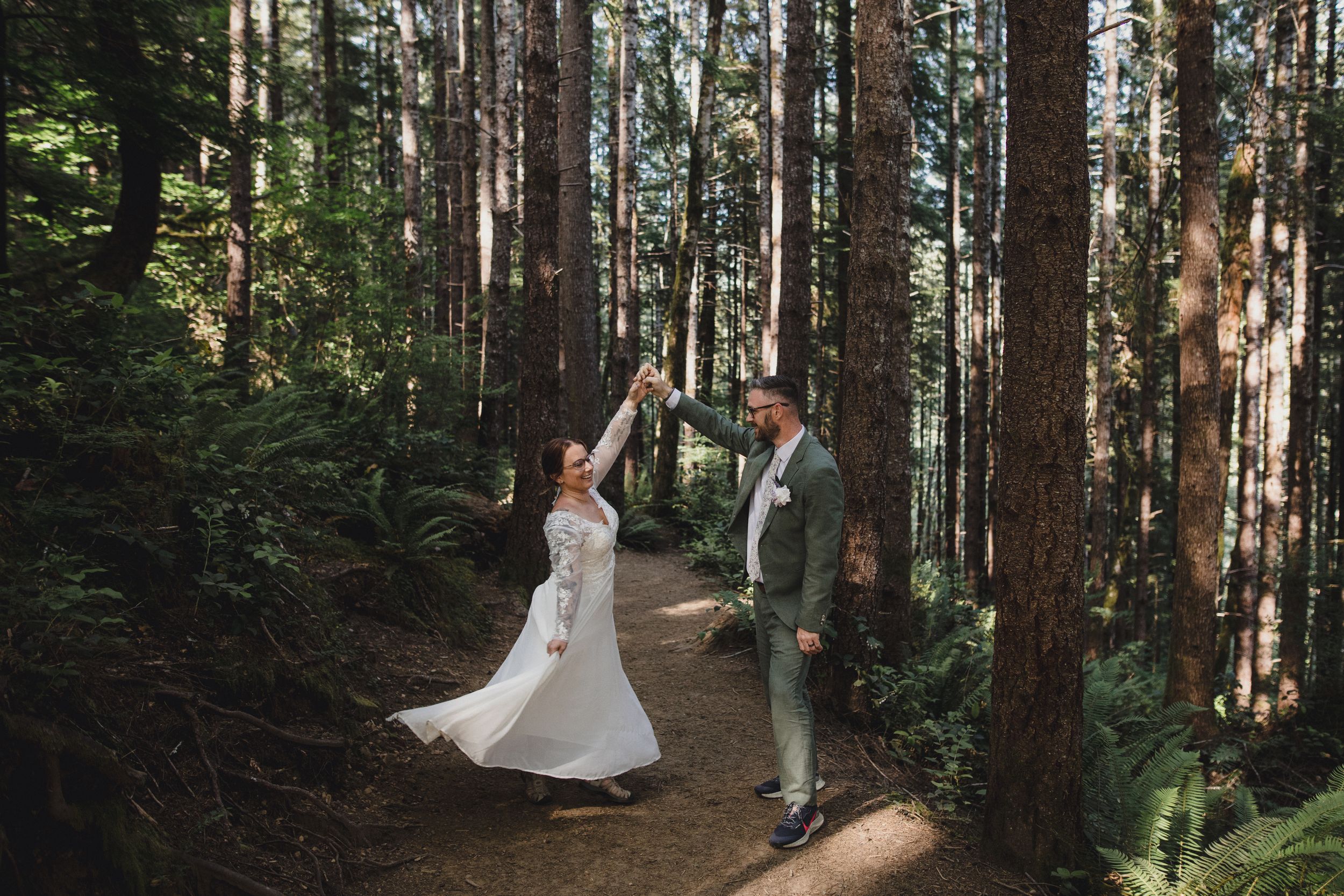 A couple dances together on a forest trail surrounded by tall evergreen trees.