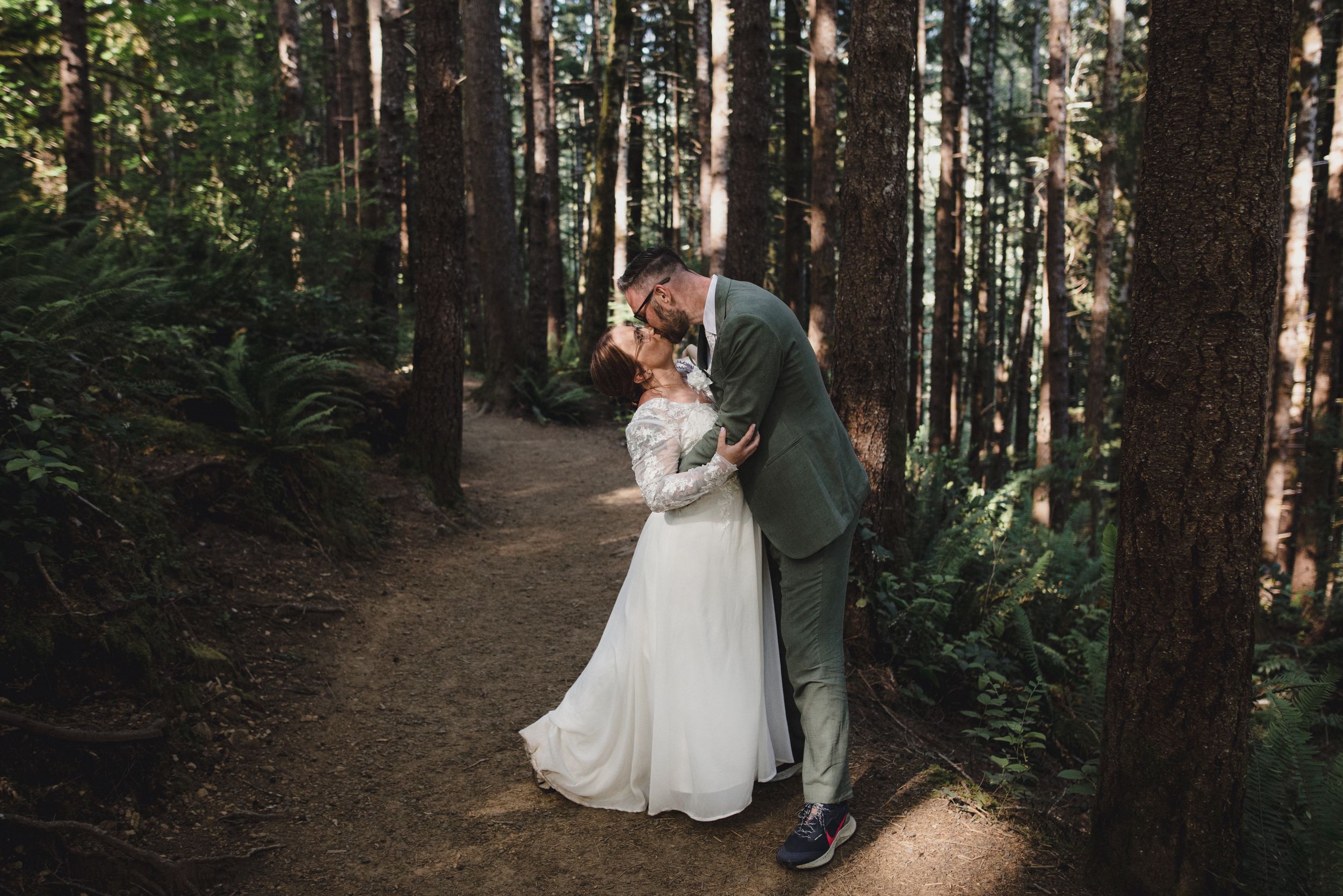 Intimate wedding moment captured on forest trail surrounded by tall evergreen trees.