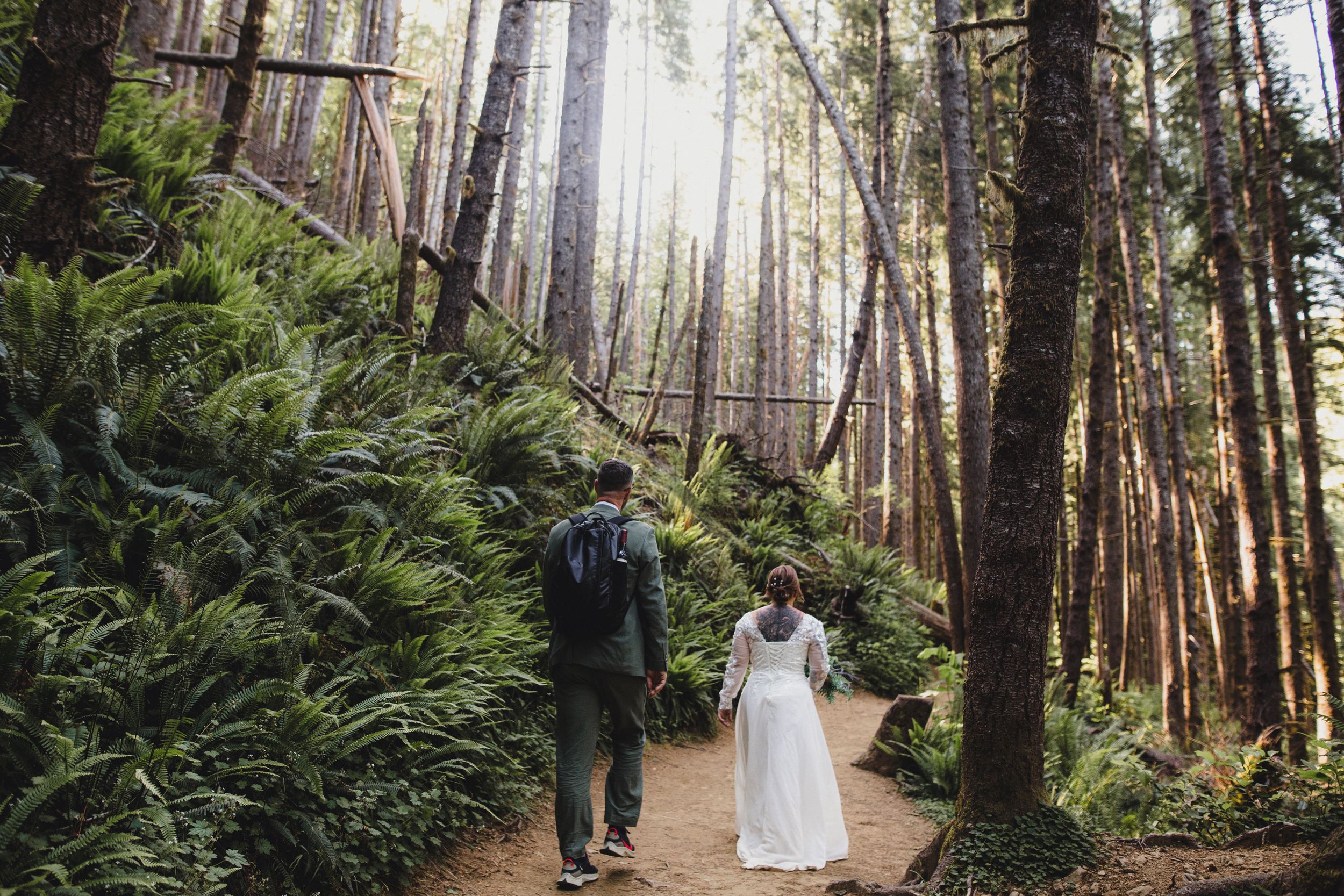 Couple walking hand in hand down a forest path surrounded by tall trees and ferns.