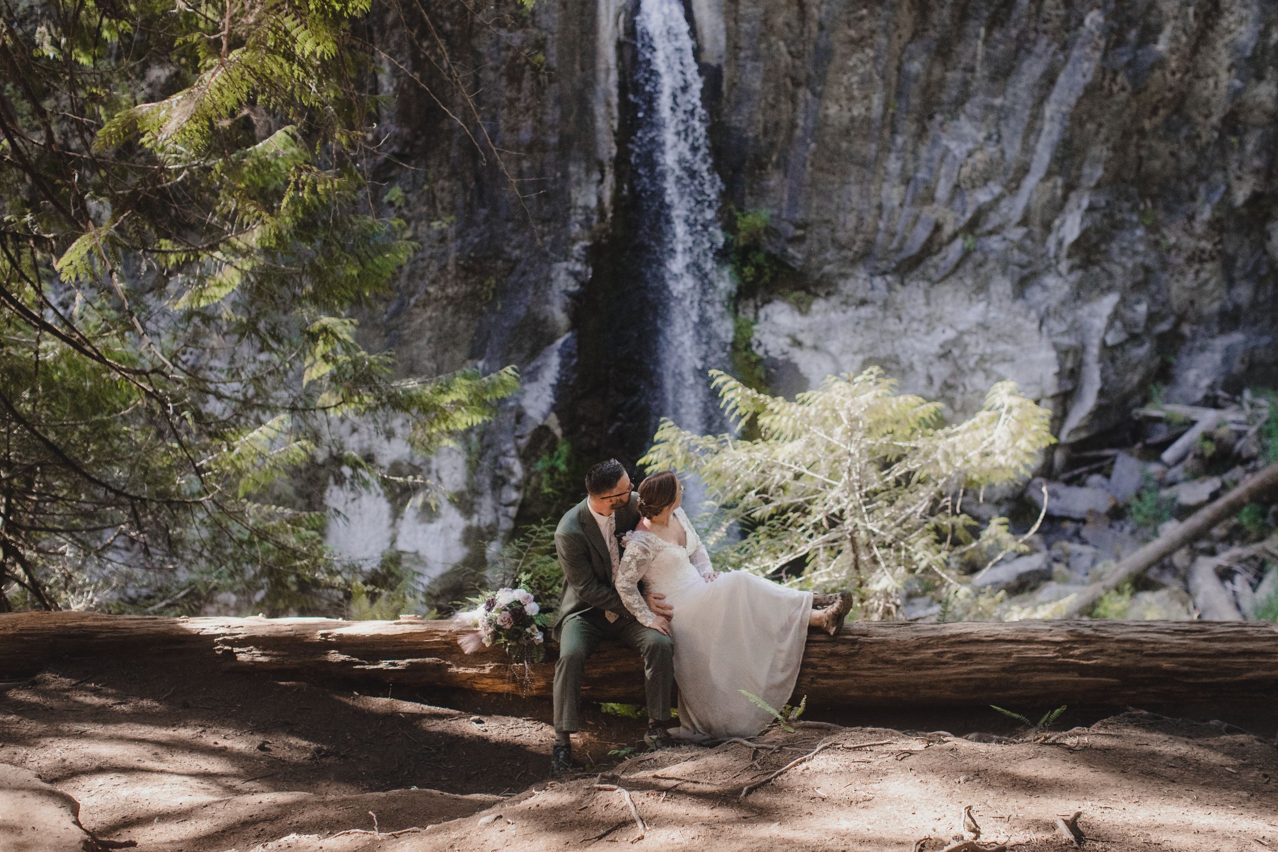 Couple resting on large log with dramatic waterfall and cliff face behind them.