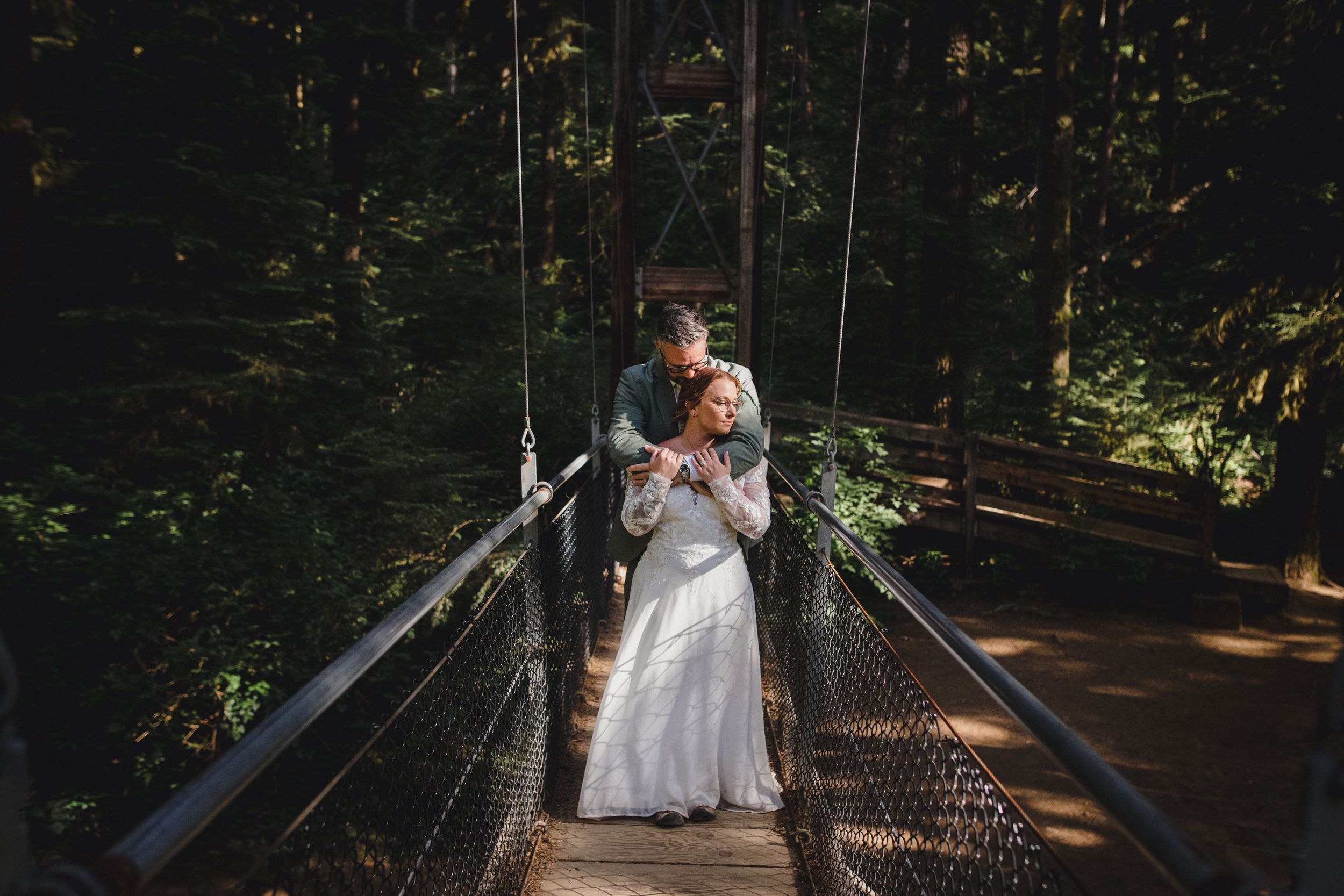 Wedding couple embraces on suspended bridge in forest setting.