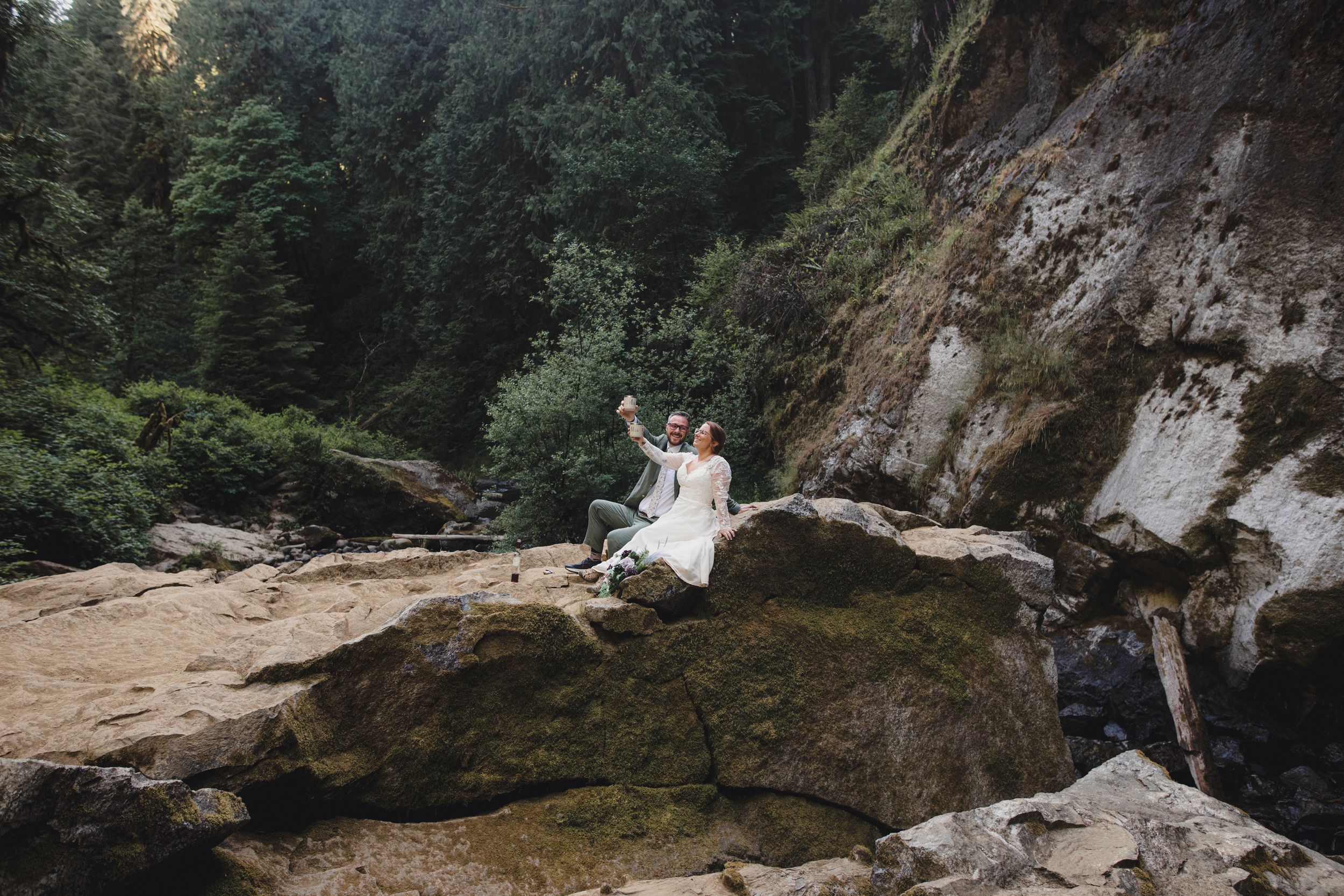 Wedding couple sitting on a dramatic rocky overlook in a forested setting.