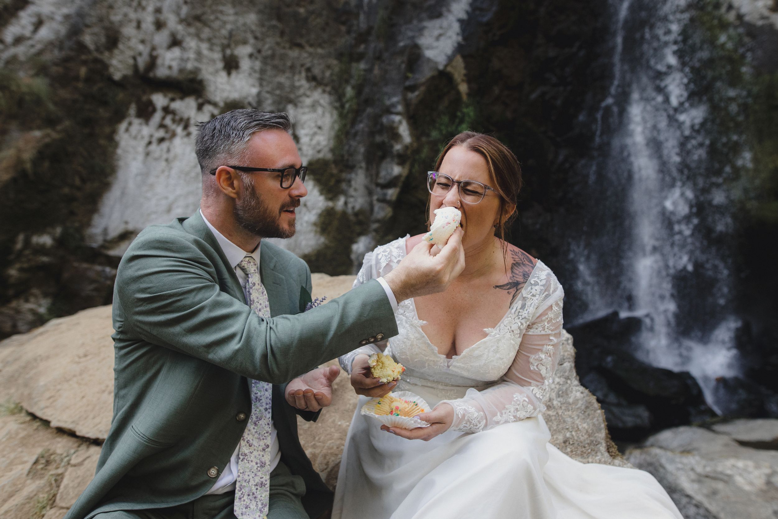 Couple playfully shares wedding cake while seated near waterfall.