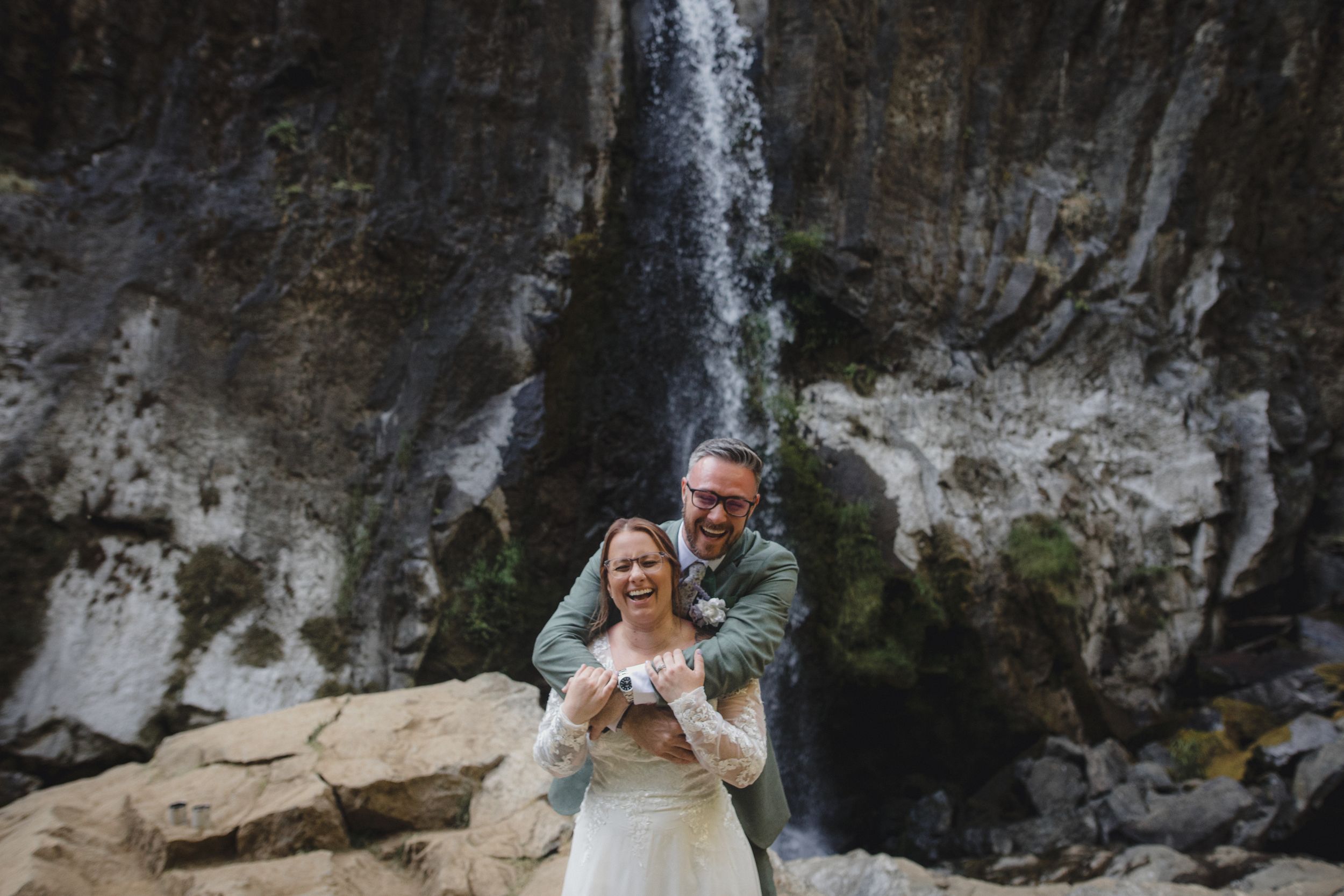 Joyful wedding couple poses in front of majestic waterfall.