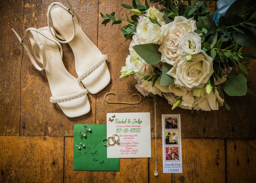 Wedding details including white bridal shoes, flowers, and invitation cards arranged on rustic wooden surface.