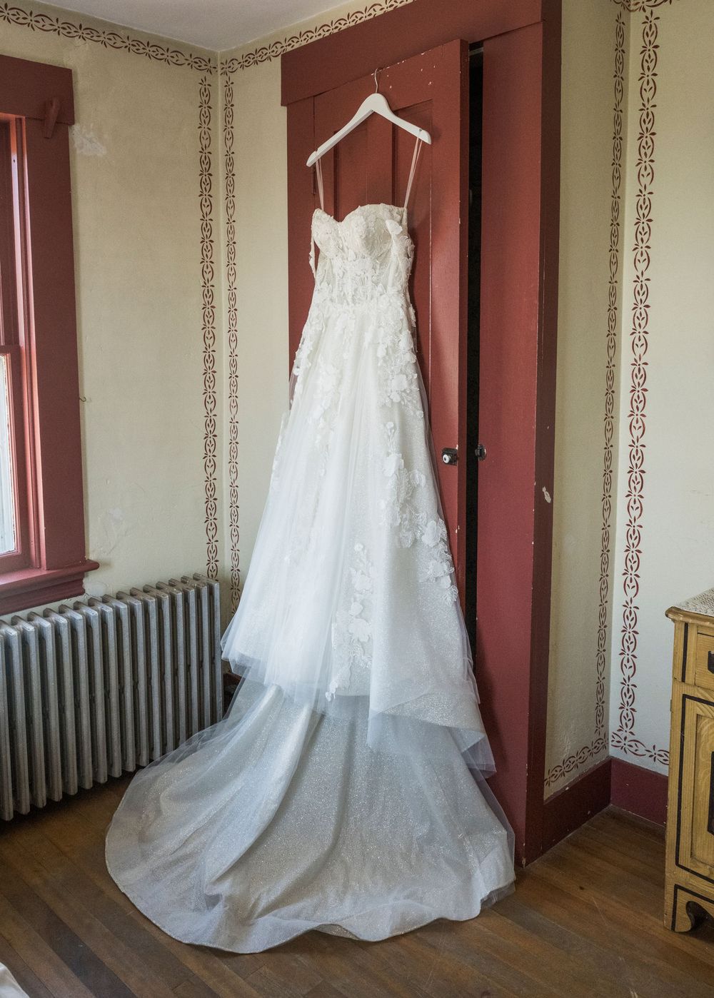 White lace wedding dress displayed on a hanger in a vintage room with ornate wallpaper and radiator.