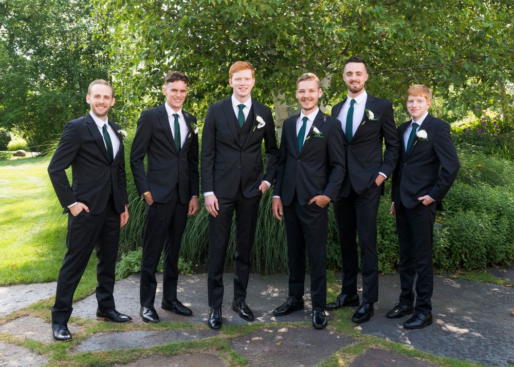 A group of groomsmen in black suits and blue ties pose together on a garden pathway.