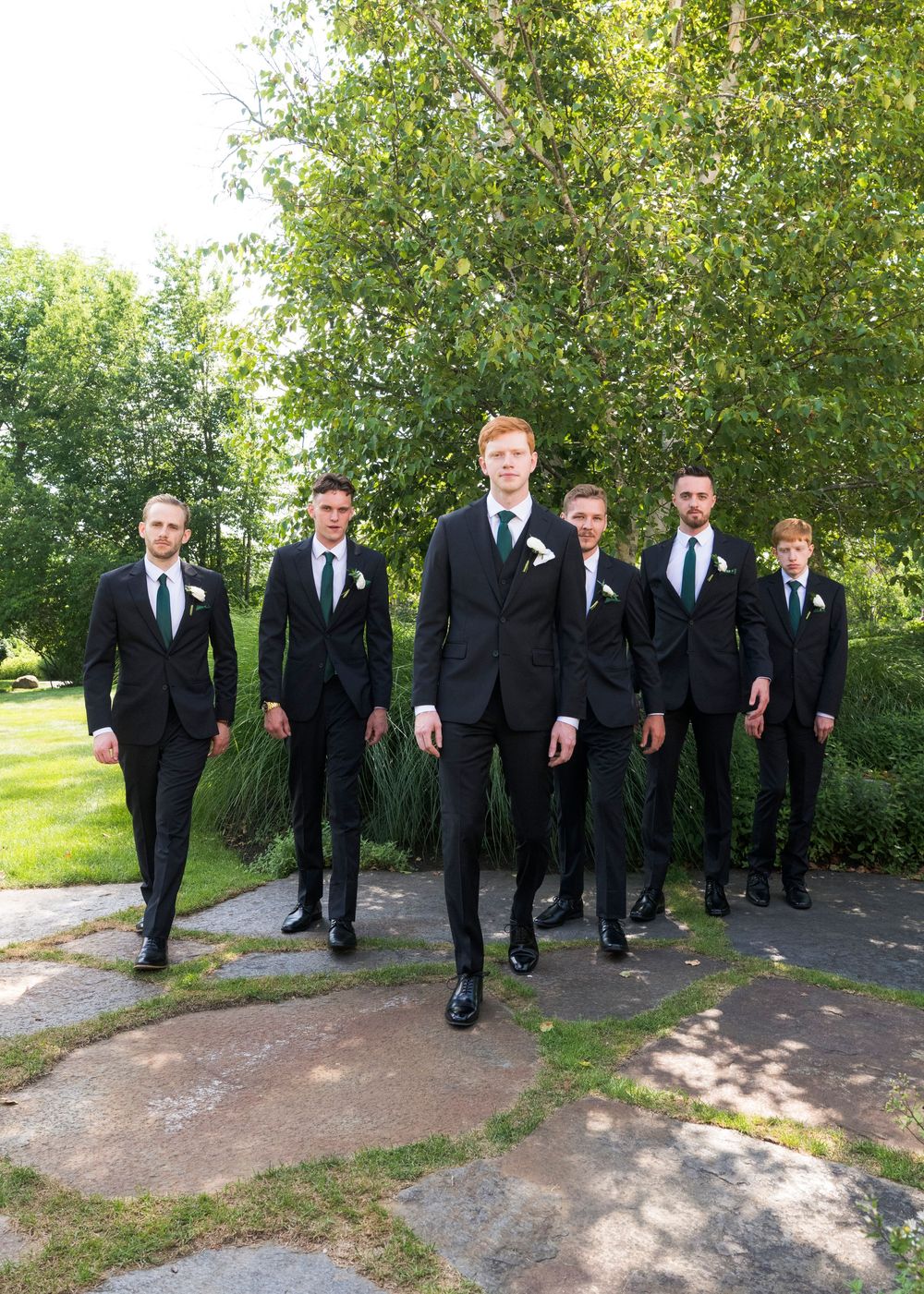 Groomsmen in black suits walk along an outdoor path lined with trees.