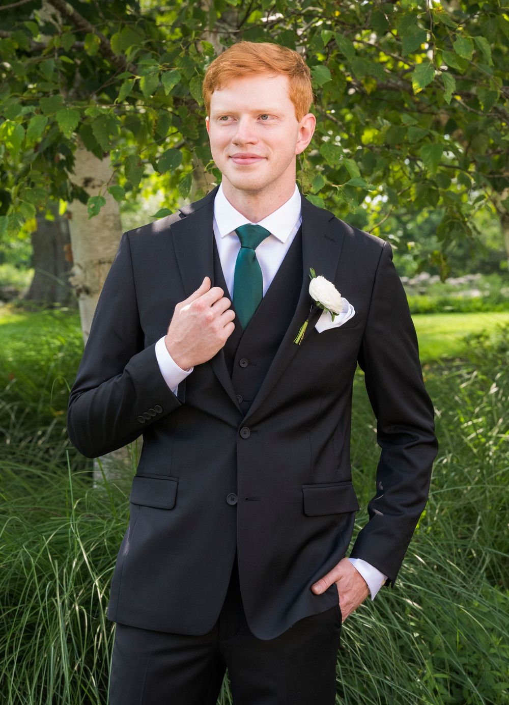 Wedding party member in a dark suit and green tie poses outdoors against green foliage.