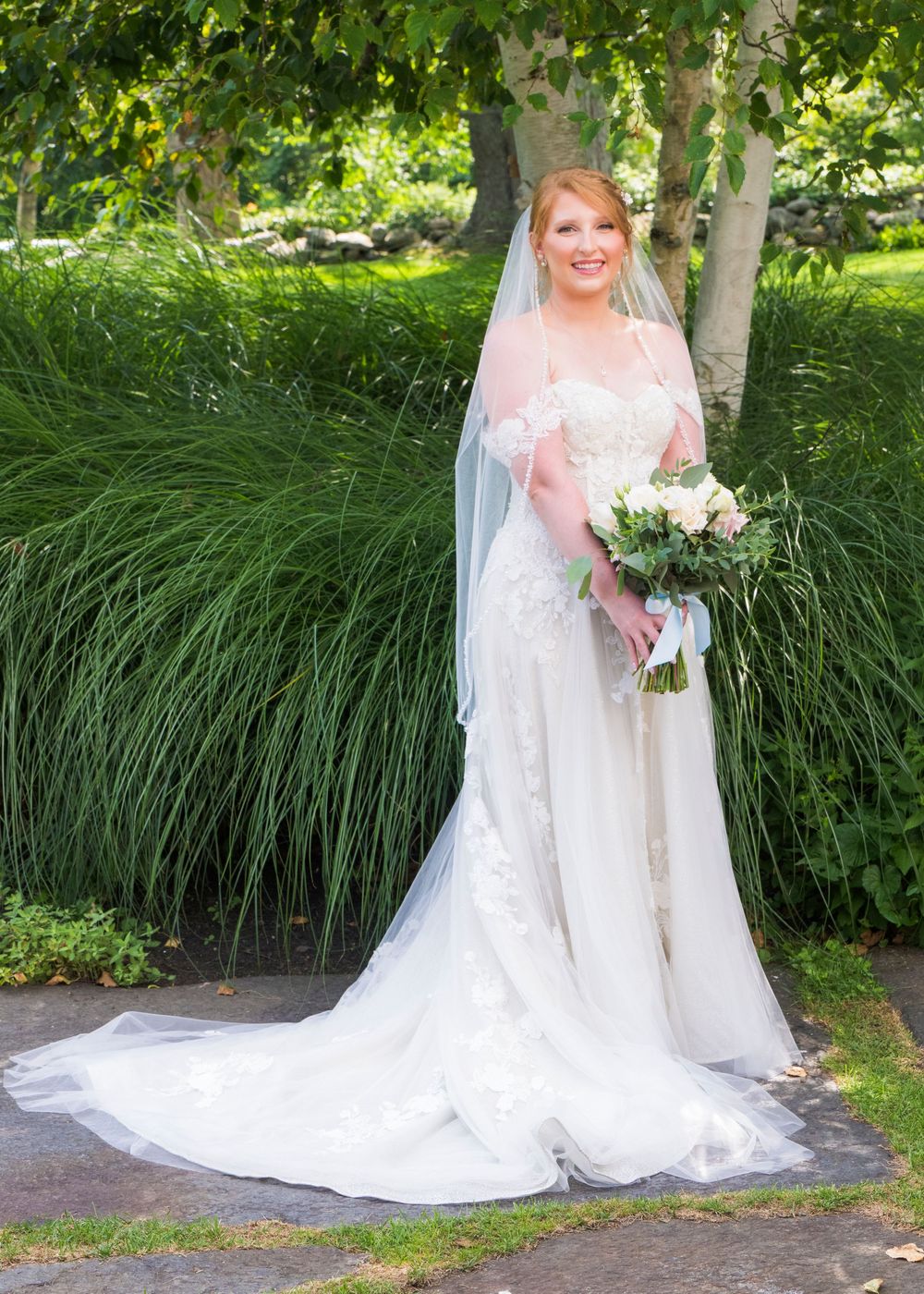 A bride in a flowing white wedding dress poses elegantly in an outdoor garden setting with tall ornamental grasses.