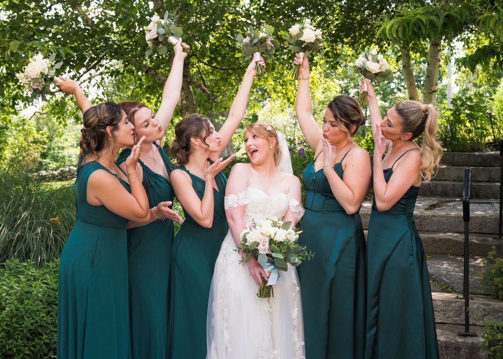 A wedding party in emerald green dresses celebrates joyfully on stone steps surrounded by lush greenery.