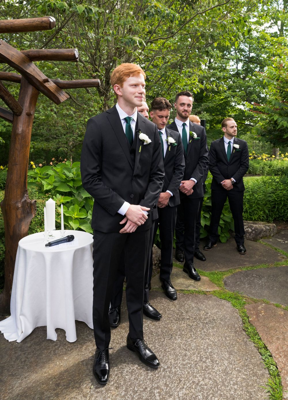 Groomsmen in black suits gather on a gravel path under a wooden pergola with white tablecloths set outside.