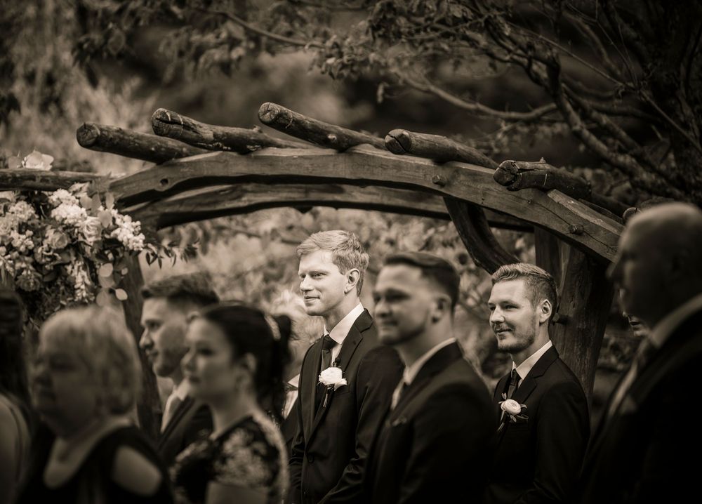 A black and white photo showing a wedding party standing together under a rustic wooden arch.