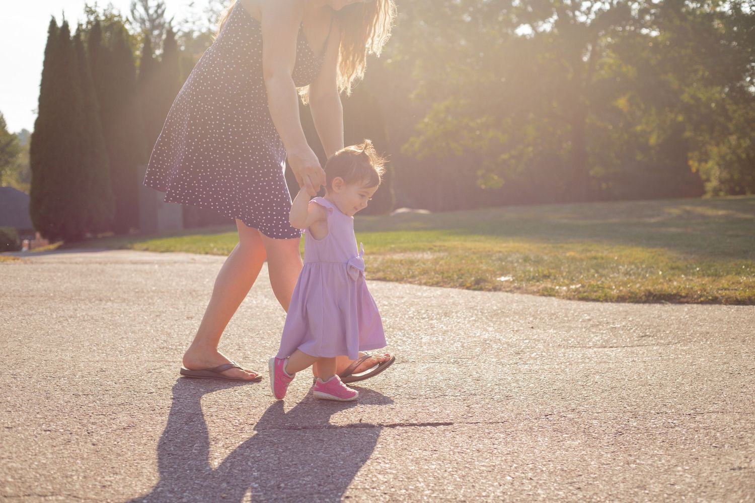 mother holding daughters hands helping her walk in the sunlight at a park