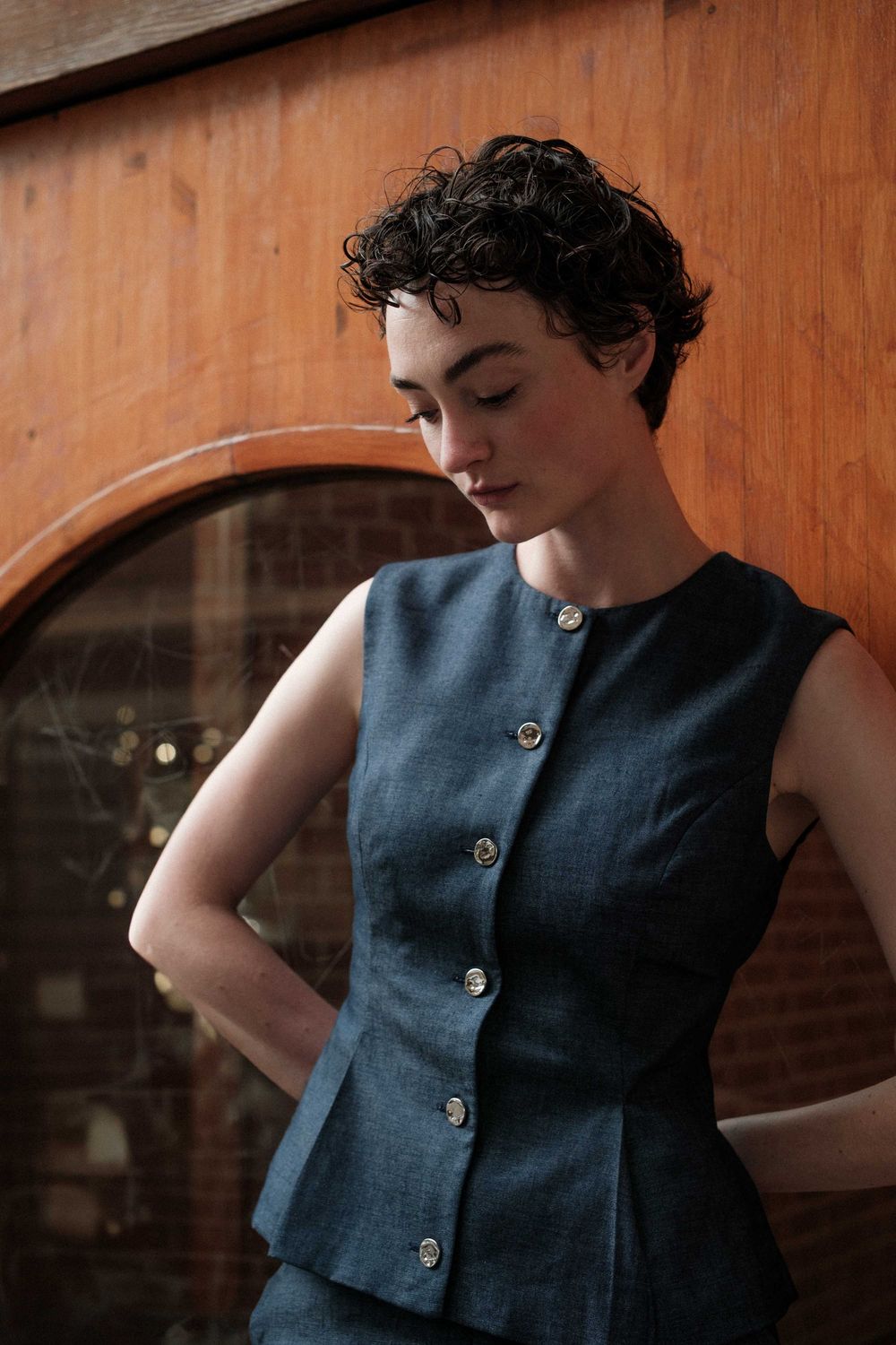 A person wearing a sleeveless blue button-up top poses in different angles against a warm wooden backdrop.