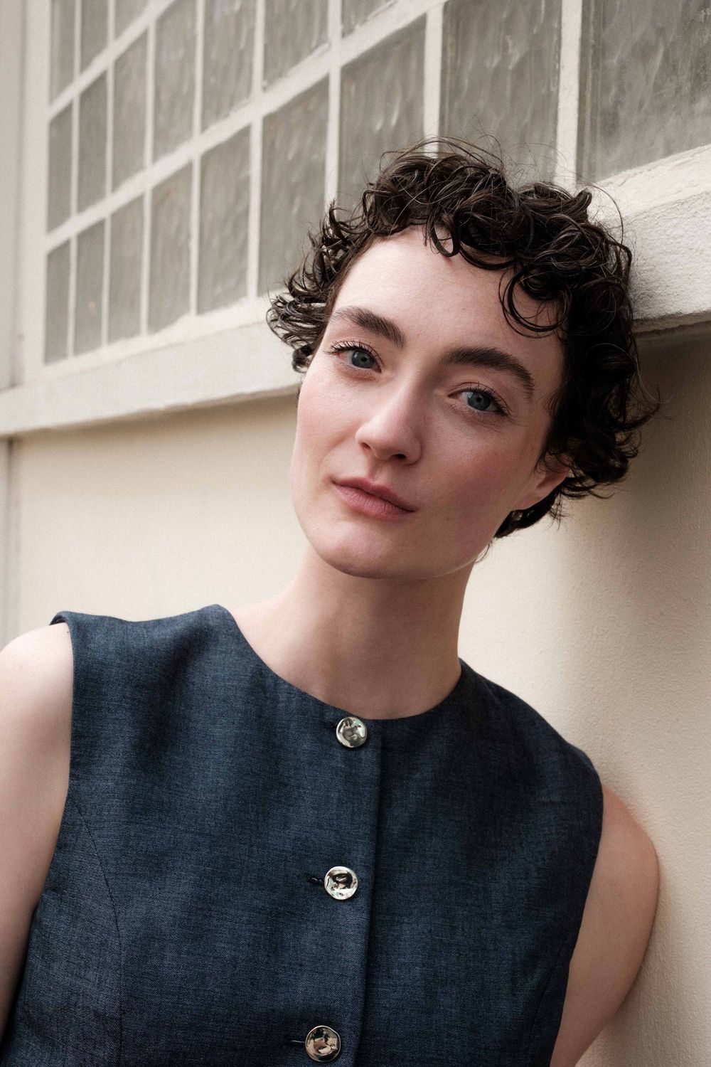 Close-up portrait against wall showing curly dark pixie haircut and sleeveless navy button-up dress.