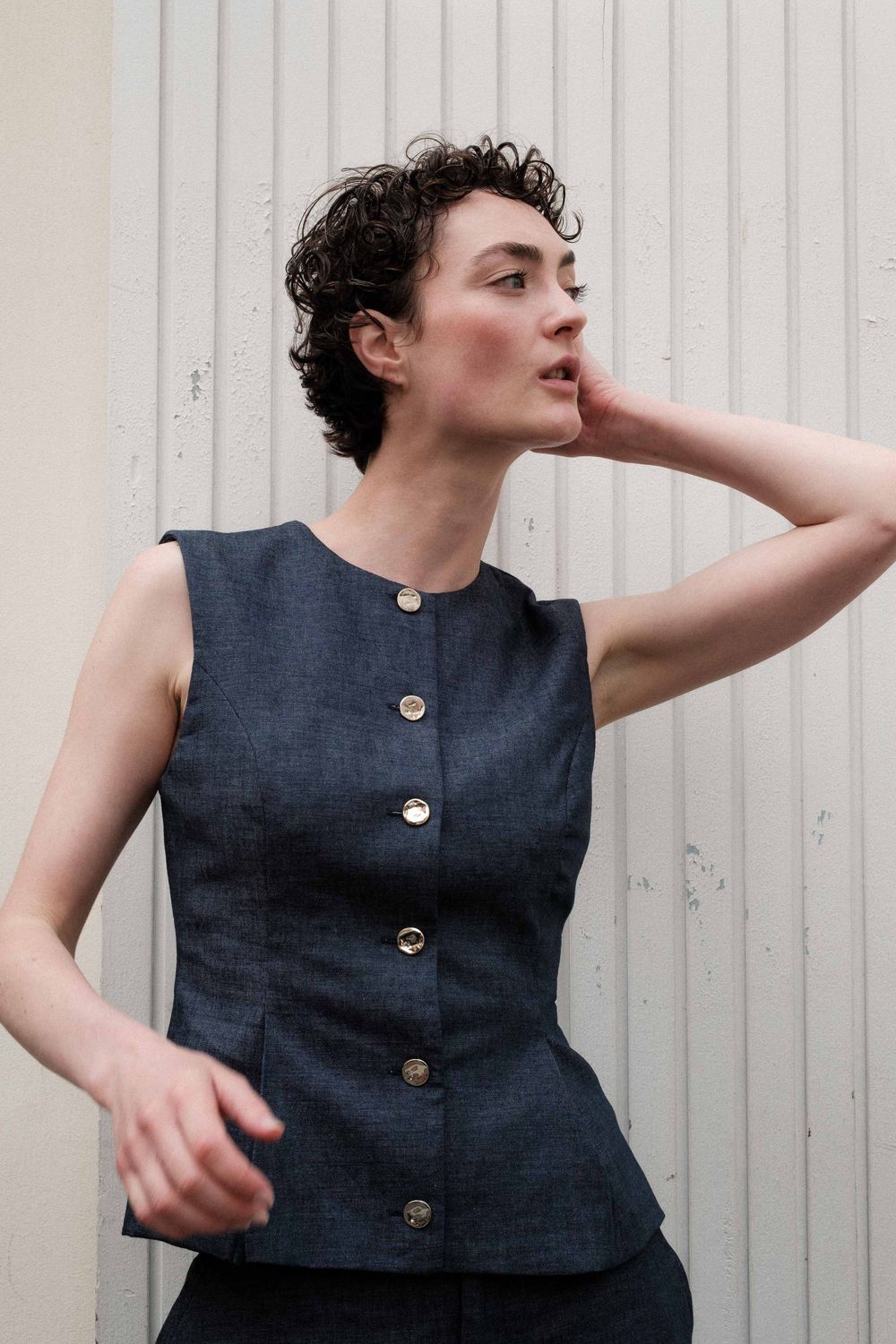Person in navy sleeveless dress with gold buttons poses playfully against white striped wall in dynamic movement series.