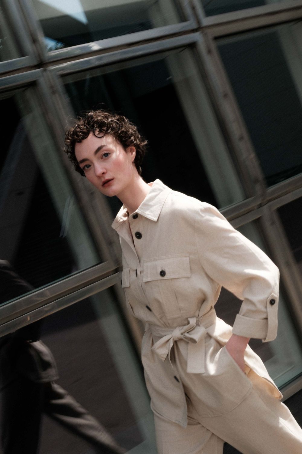 Model in cream-colored belted utility jacket poses against window backdrop during fashion photoshoot.