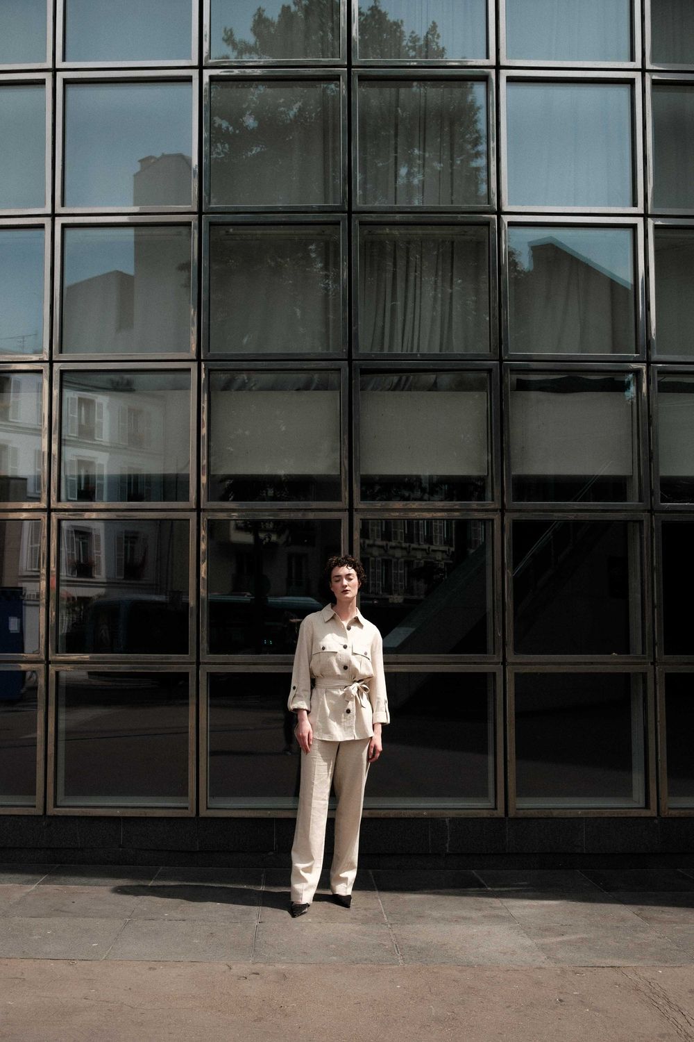 Person wearing a beige suit stands in front of a modern glass building facade with geometric window panels.