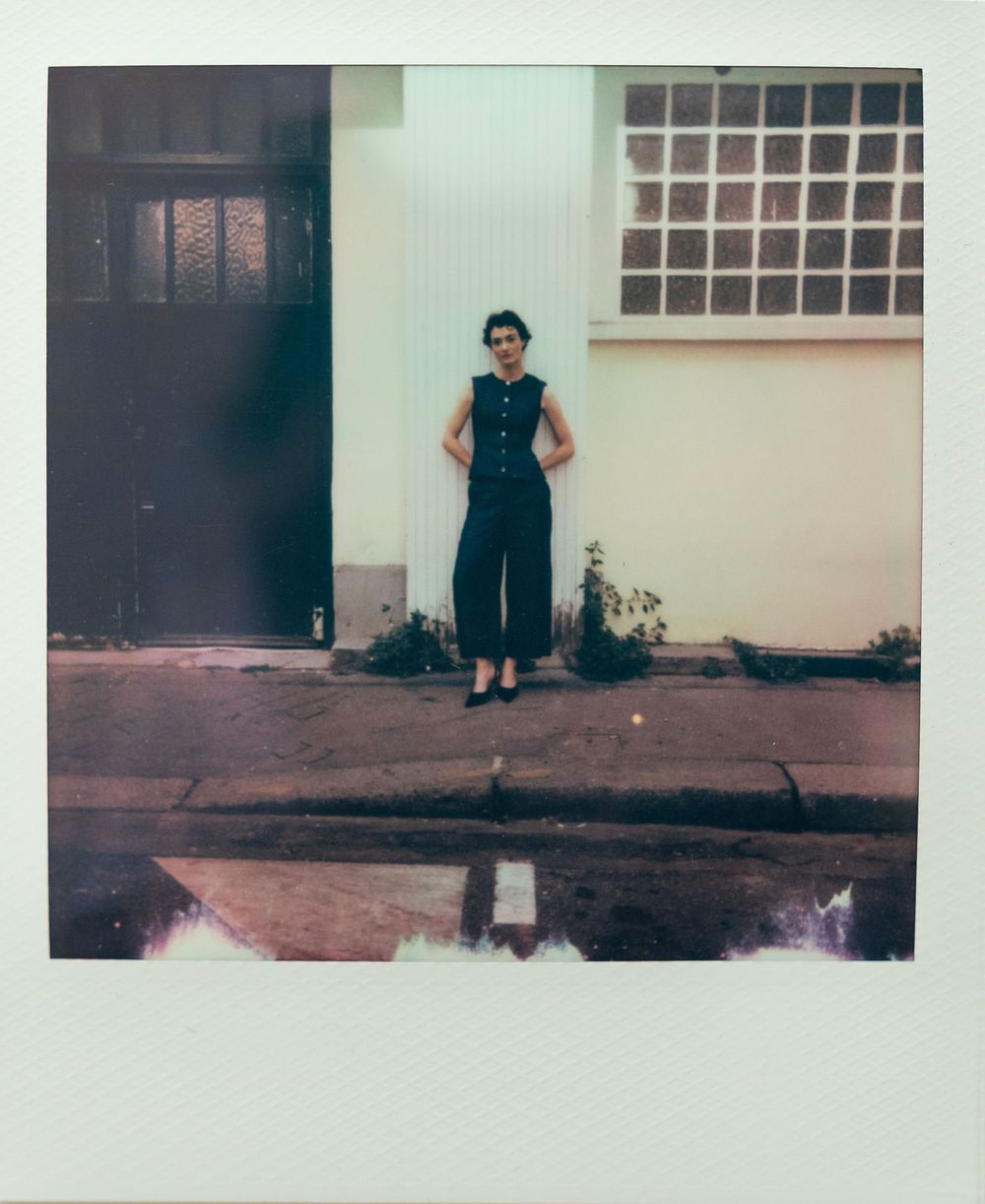 Vintage polaroid photograph showing a person in a dark dress standing against a light-colored wall with a window and door.