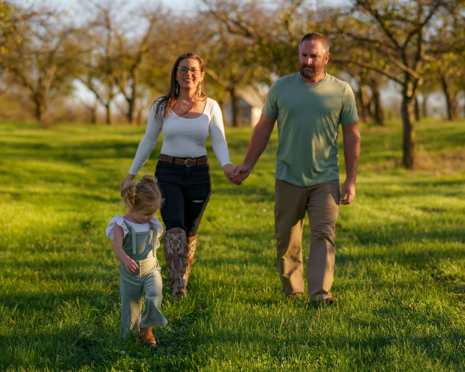 Family of three walking hand-in-hand through a sunny green orchard on a warm spring day.