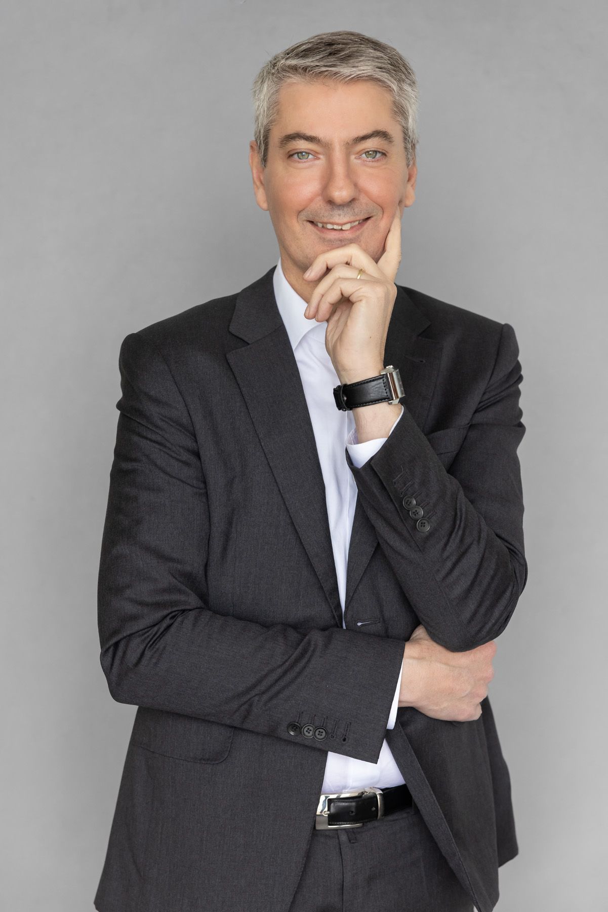 A middle aged professional in a dark suit and white shirt poses thoughtfully with hand on chin against gray background.