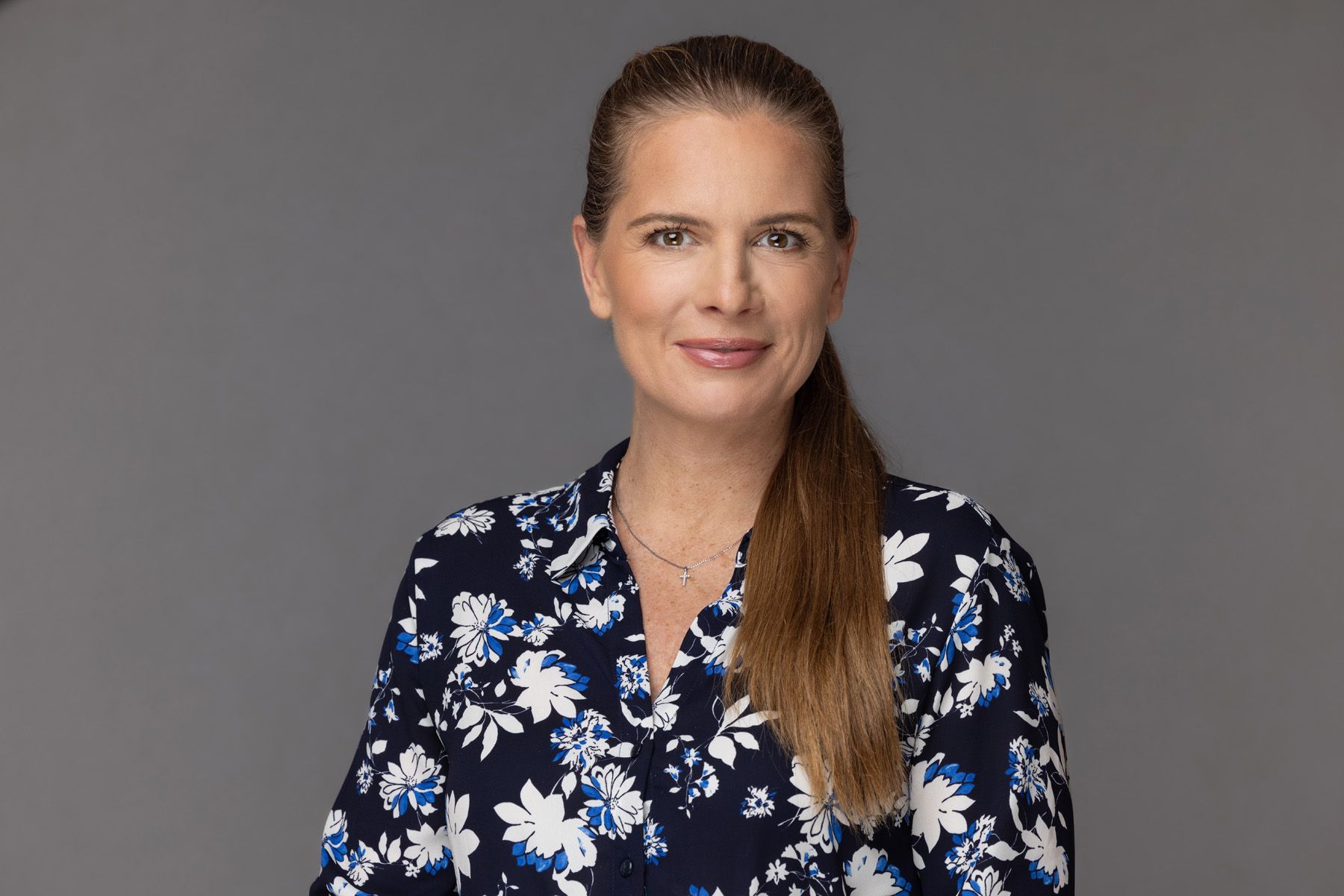 Professional headshot against gray background featuring long blonde hair and navy floral print blouse.