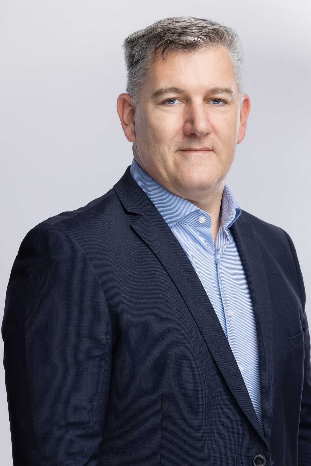Professional headshot of person in a dark suit jacket and light blue button-down shirt against gray backdrop.