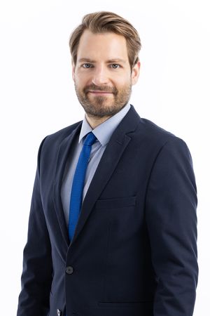 Professional headshot of person in dark suit with blue tie posing against light background.