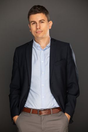 Professional portrait of someone in a dark suit jacket, light blue button-down shirt and khaki pants against grey backdrop.