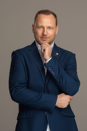 Professional business portrait of a person in a navy suit with hand on chin against gray background.
