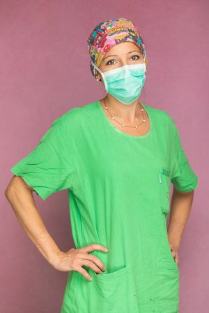 Healthcare worker in green scrubs, surgical mask and colorful floral scrub cap poses in front of pink background.