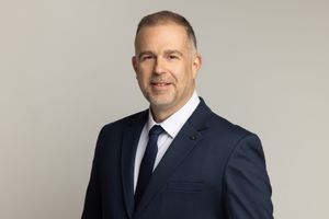 Professional portrait of a business executive in a navy suit and tie against a light gray background.