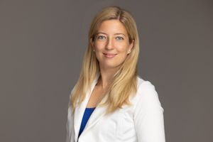 Professional headshot of a person in a white blazer and blue top against a neutral grey background.