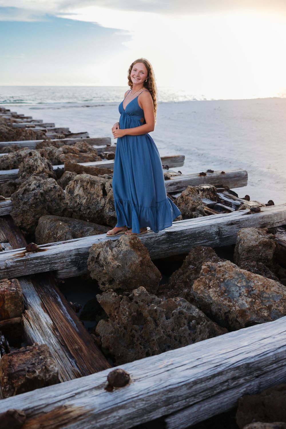 A flowing blue maxi dress contrasts beautifully against rocky coastal beach terrain at sunset.