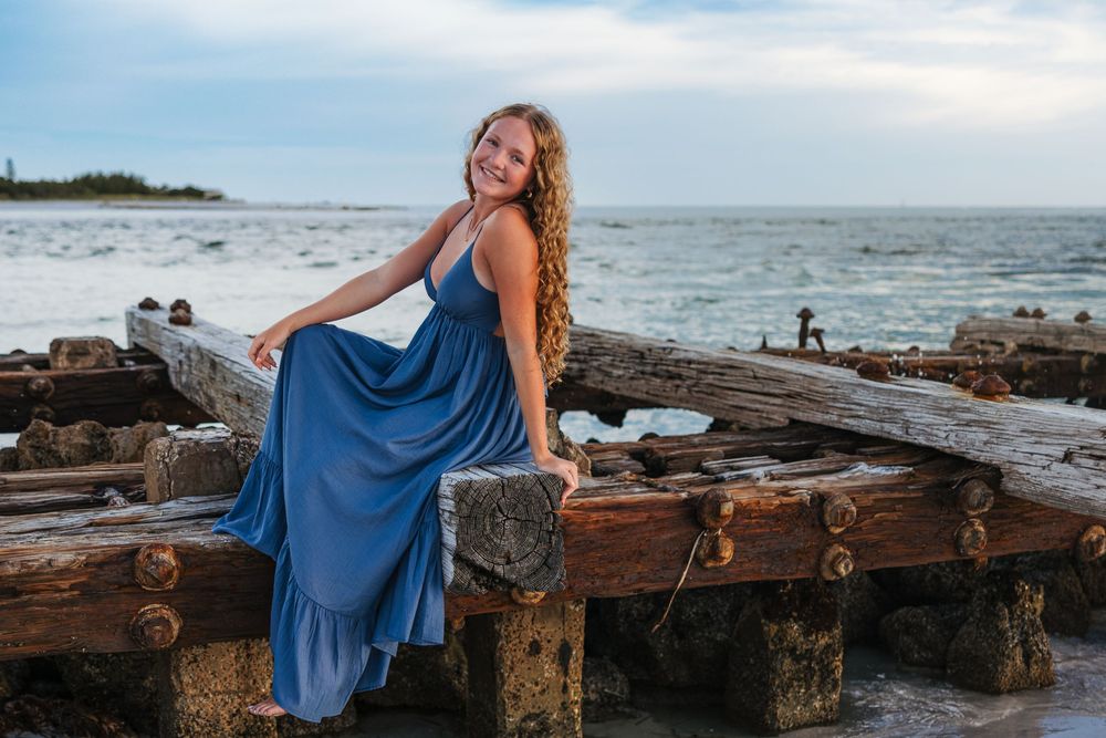 A figure in a flowing blue dress poses on rustic wooden beams along a rocky beach at sunset.