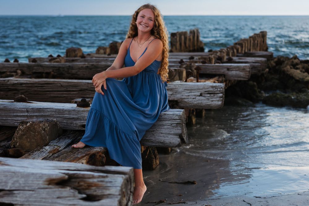A breezy blue sundress catches the light while seated on weathered driftwood by the ocean.